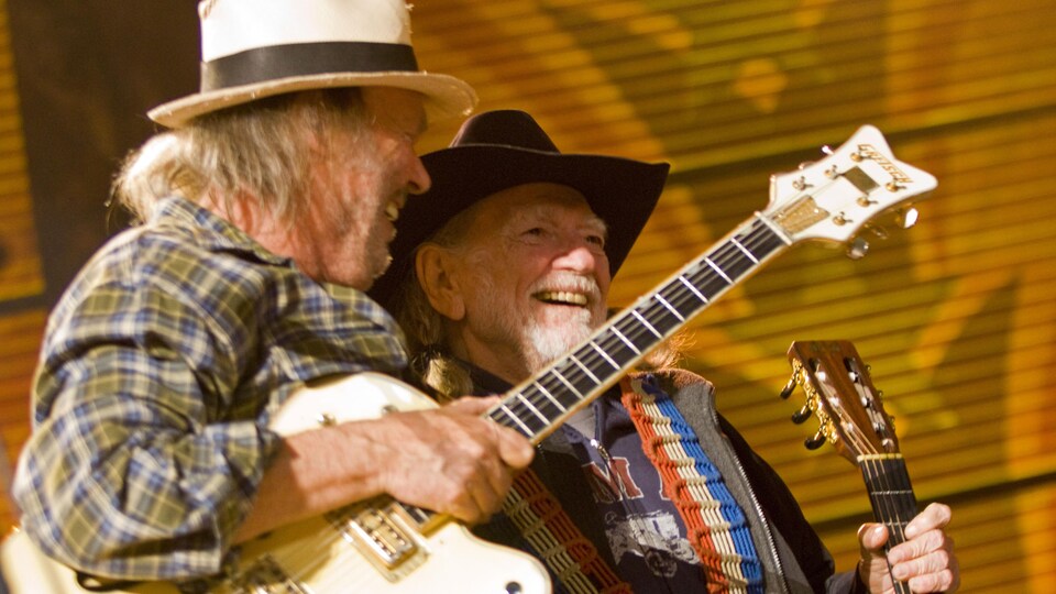 Deux hommes portant des chapeaux et tenant des guitares sourient sur scène. 