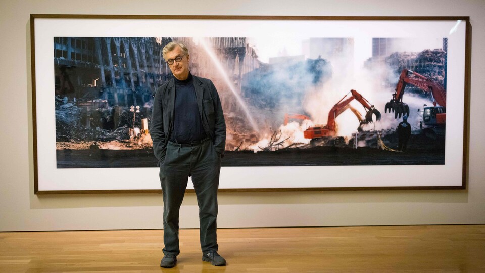 L'homme se tient devant l'une de ses photos, main dans les poches, et penche la tête comme s'il tentait d'éviter le jet d'eau qu'il y a dans la photo derrière lui.