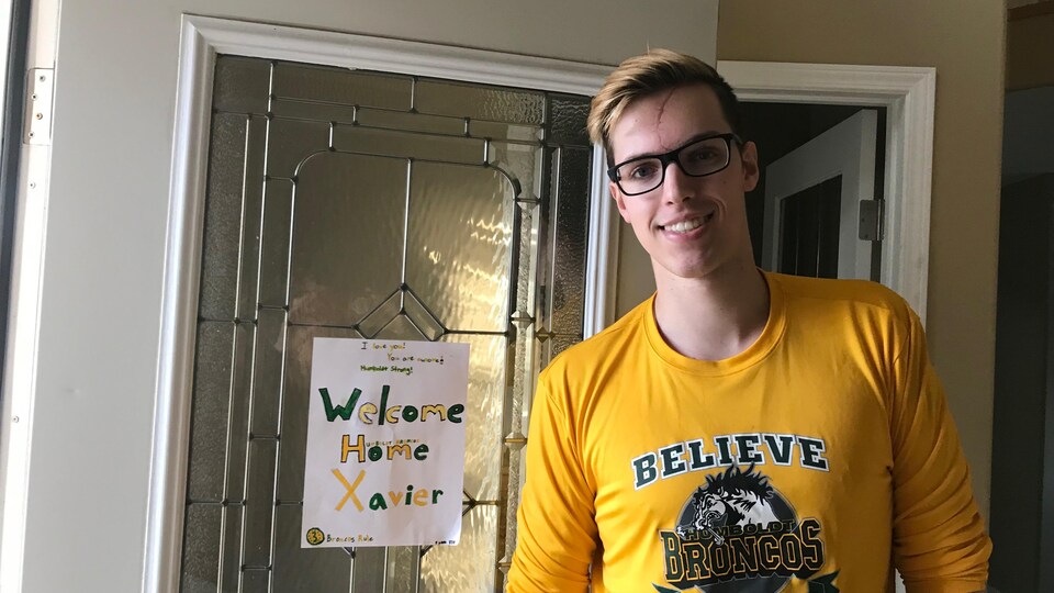 Un jeune homme portant le chandail aux couleurs des Broncos de Humboldt devant la porte d'entrée de sa maison avec une pancarte pour lui souhaiter un bon retour à la maison