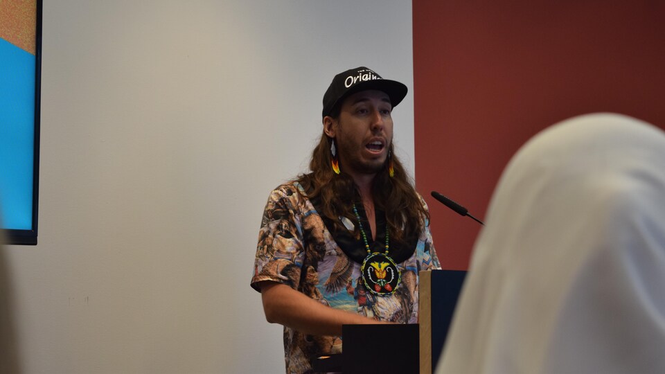 Un homme portant une casquette et un collier autochtone parle devant un public dans une salle.