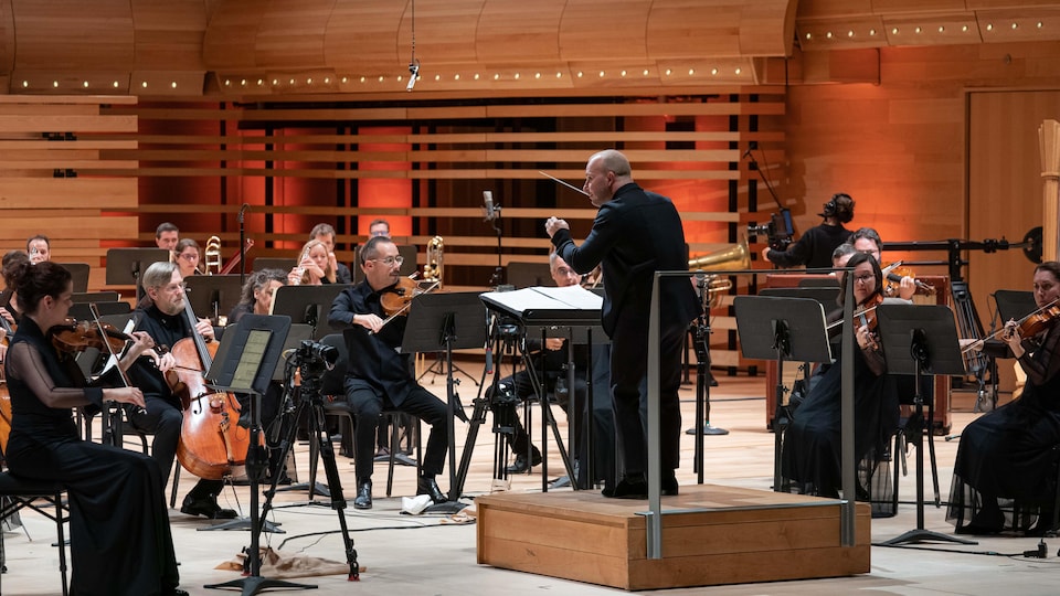 Un chef d'orchestre sur son podium dirige une douzaine d'instrumentiste dans une salle de concert.
