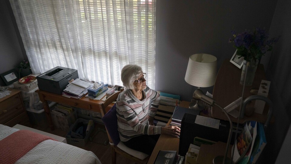 Une dame à l'ordinateur dans la pénombre de sa chambre où l'on voit une partie de son lit, des livres, une imprimante.