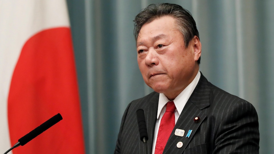 Une photo de Yoshitaka Sakurada pendant une conférence de presse. Il porte un veston noir aux fines rayures verticales, une chemise blanche et une cravate rouge. Un drapeau du Japon est visible derrière lui.