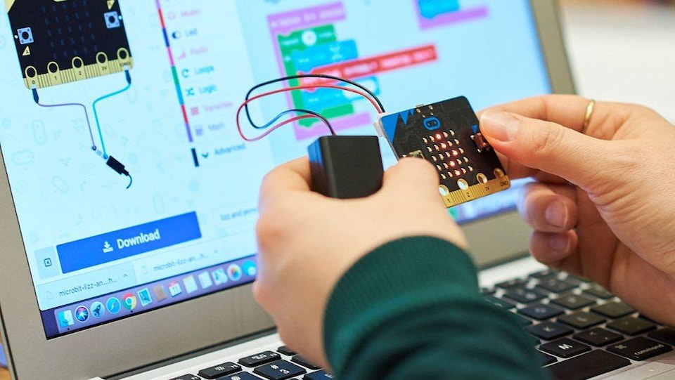 Zoom sur deux mains qui tiennent une pièce informatique raccrochée à un boitier par deux fils électriques. Derrière se trouve un écran d'ordinateur.