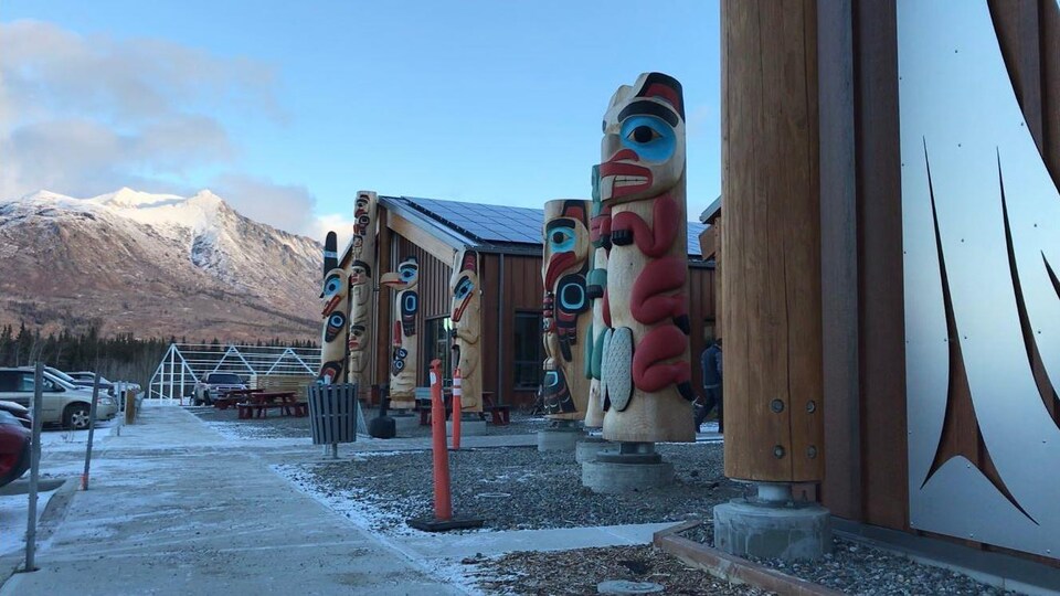 Des totems devant un édifice avec des montagne enneigées en arrière-plan.