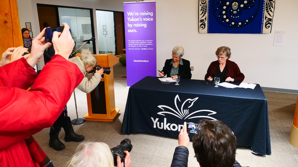 Plan large de Jeanne Beaudoin et de Janet Welch signant un document à une table devant plusieurs photographes.