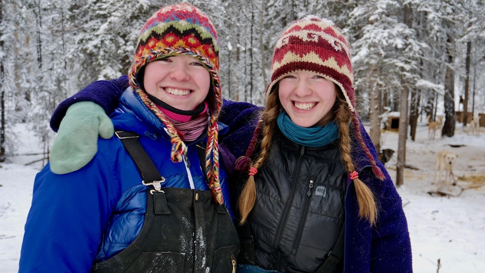 Deux jeunes filles de 18 ans qui se ressemblent sourient à la caméra. Elles sont dehors, dans la neige. Celle de droite a le bras droit passé sur les épaules de celle de gauche.