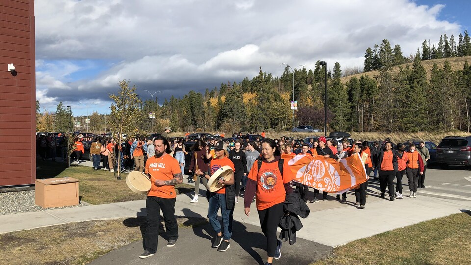 Un groupe de jeunes qui marchent et sont vêtus de chandails orange, ils tiennent des tambours.