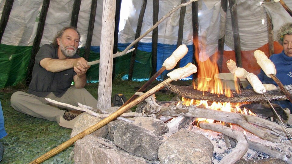 Yv Bonnier-Viger autour d'un feu qui cuit un morceau de pain banique sur un bâton de bois.