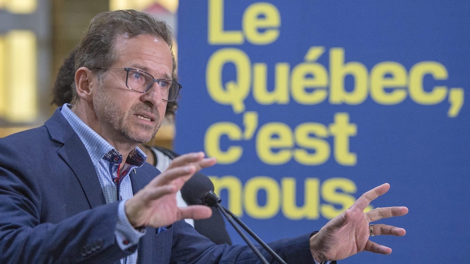 Yves-Francois Blanchet parle dans un micro, devant une affiche sur laquelle on peut lire « Le Québec, c'est nous ».