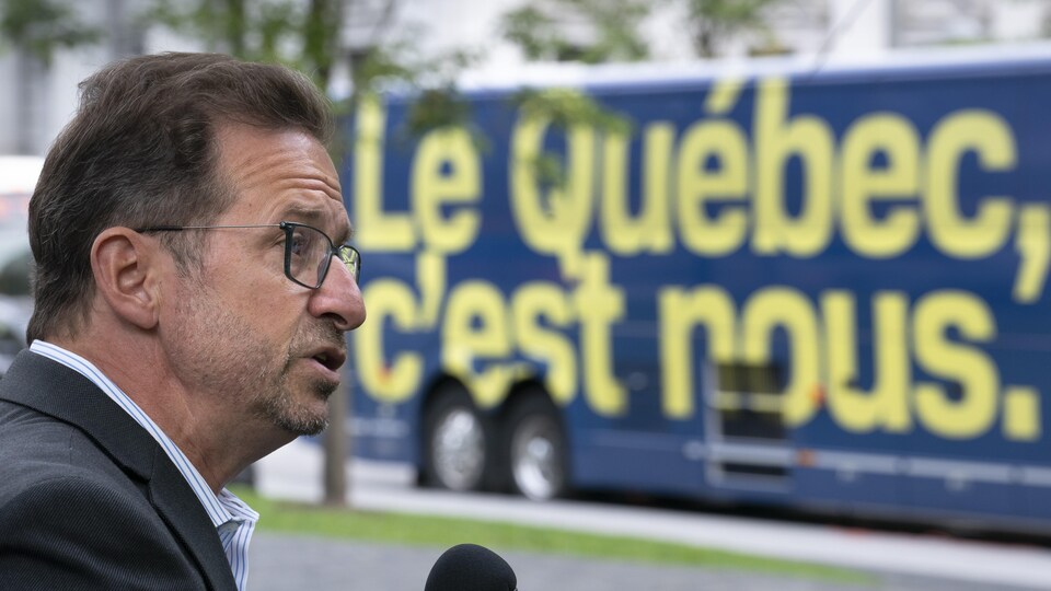 Yves-François Blanchet est de profil et parle au micro. Derrière lui, l'autobus du Bloc québécois est stationné.