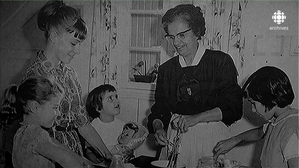 Photo en noir et blanc d'Yvonne Maisonneuve, cuisinant aux côtés d'une jeune fille et de trois enfants