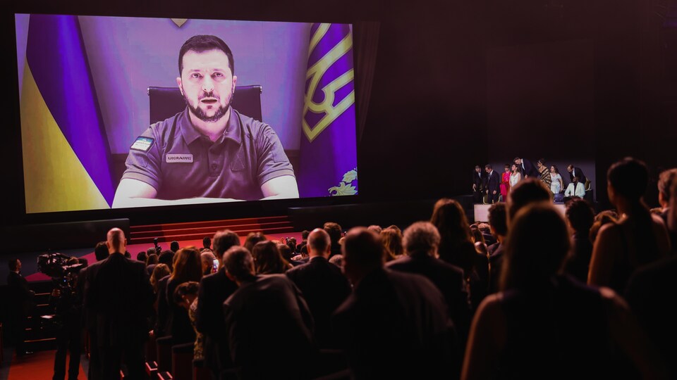 Des gens se lèvent de leurs sièges dans un amphithéâtre pendant la projection d'un message vidéo du président ukrainien. 