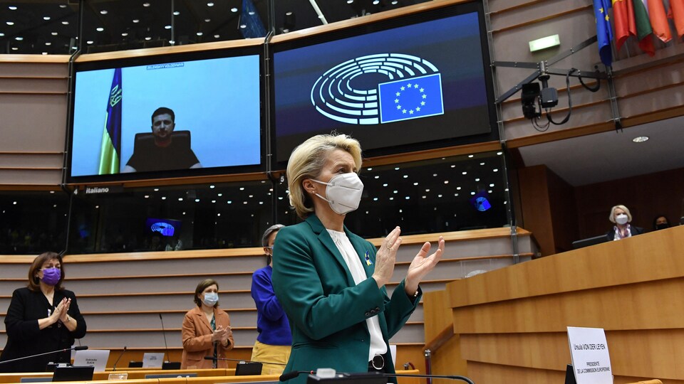 Ursula von der Leyen, debout, applaudit, comme d'autres personnes près d'elle, dans l'enceinte du Parlement européen. M. Zelensky est visible sur un écran. 