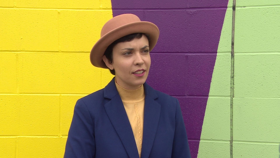 L'artiste visuelle Zoé Fortier qui porte un chapeau devant dehors devant une peinture murale colorée.