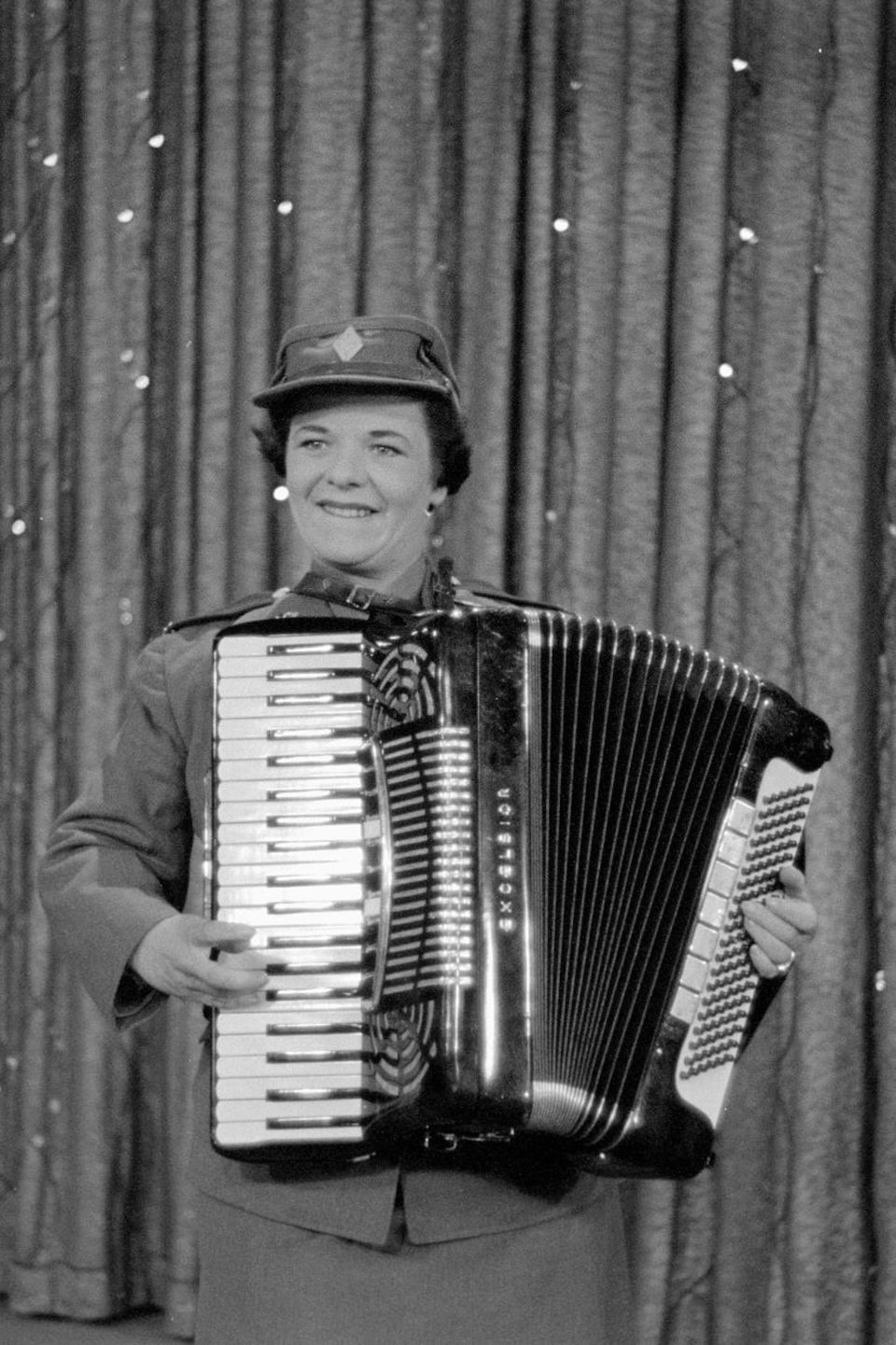 Une accordéoniste en uniforme joue de son instrument en souriant sur une scène avec un rideau pailleté.