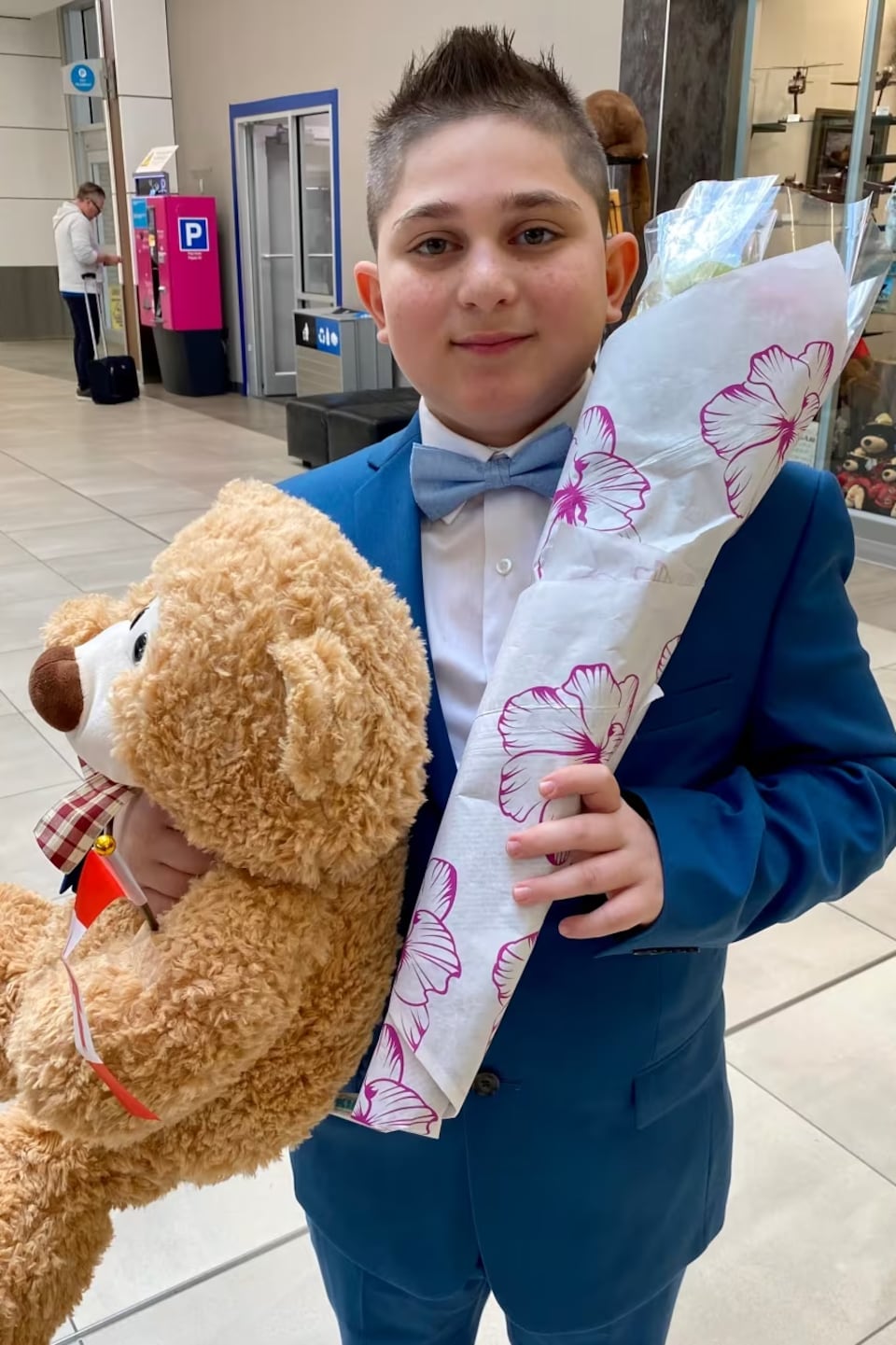 Adnan Kharsa porte un costume bleu et un nœud papillon. Dans ses mains, il tient un bouquet de fleurs et un ours en peluche.