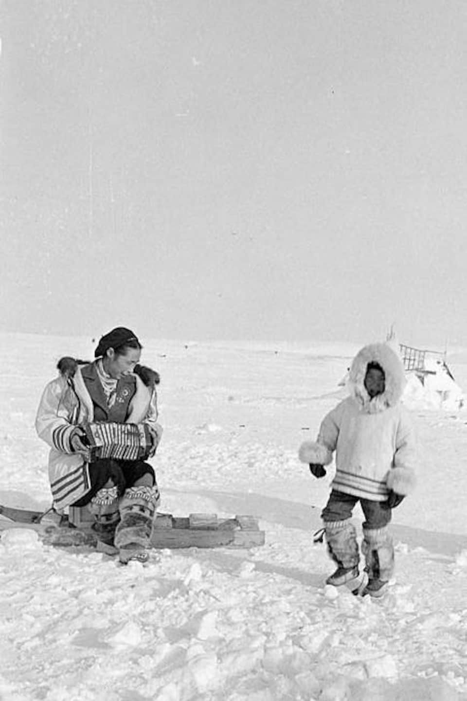 Photo d'archive d'Allie Salluviniq en tenue traditionnelle d'hiver est en train de danser devant d'autres personnes chaudement habillées, sur sa droite Edith Patsau joue de l'accordéon, sur la banquise dans le nord de l’île d’Ellesmere, en mars 1956.