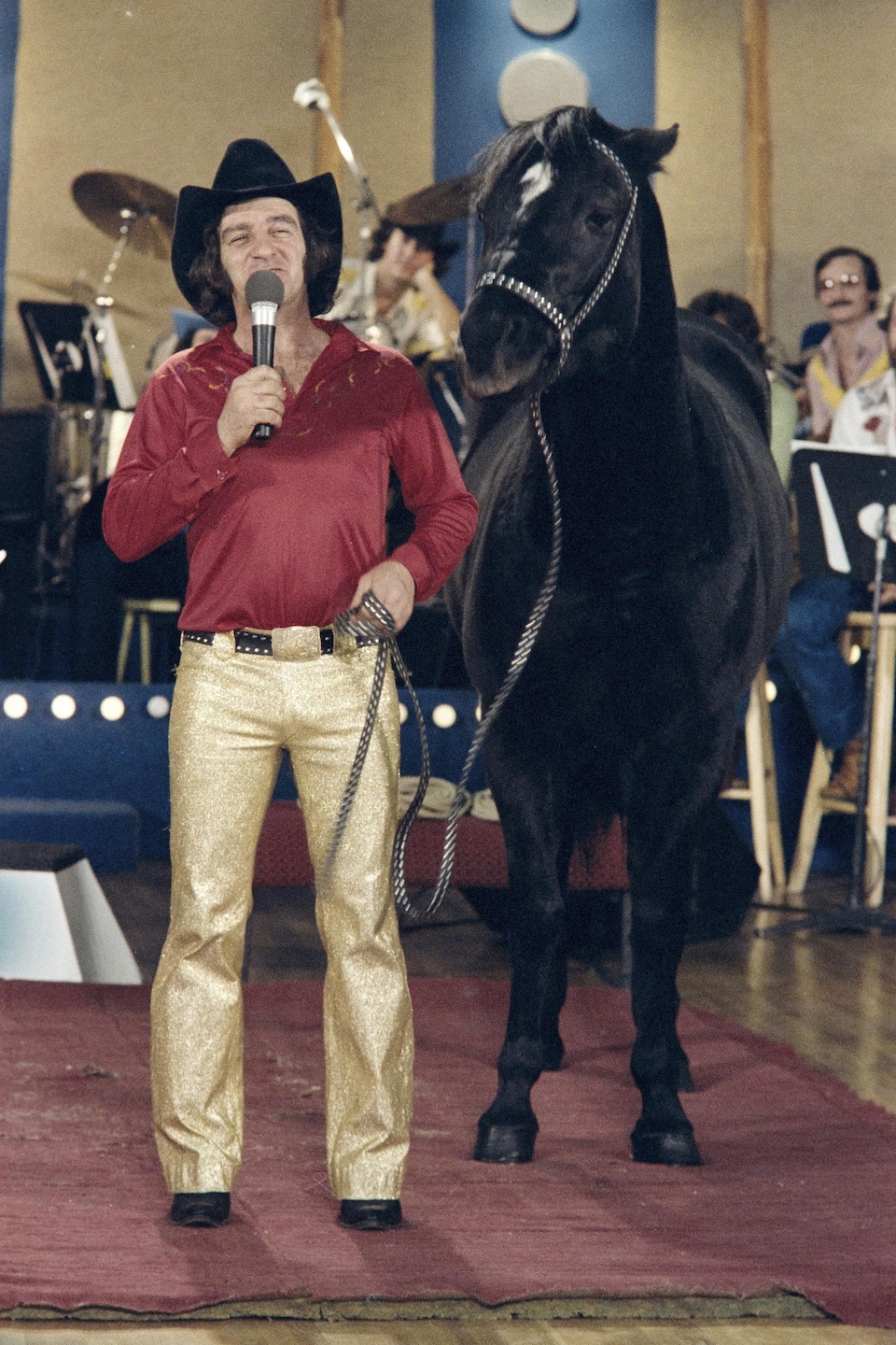 Micro à la main, le cowboy Bucksin tient son cheval par une longe sur le plateau de l'émission, avec les musiciens en arrière-plan.