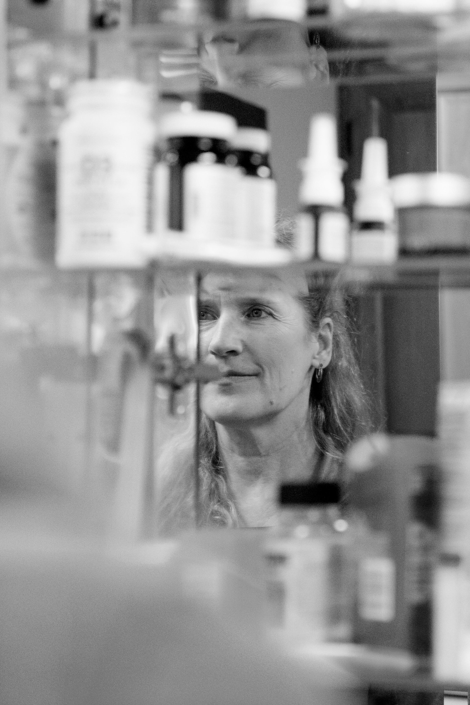 Une femme devant un miroir, avec des flacons de médicaments sur des tablettes.