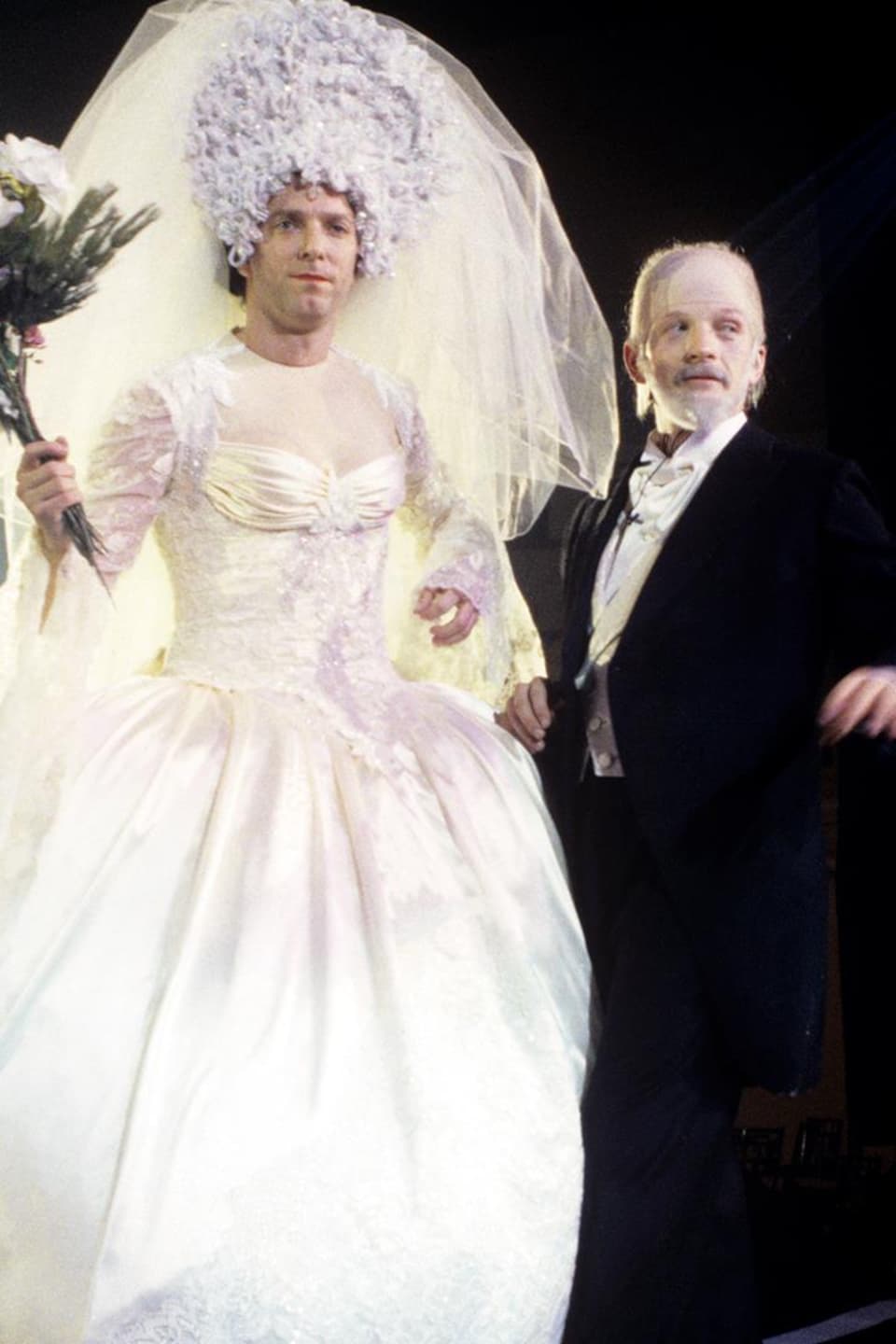 Vêtue d'une robe de mariée, Céline Dion (Patrice L'Écuyer) est debout, au bras de René Angelil (André-Philippe Gagnon).