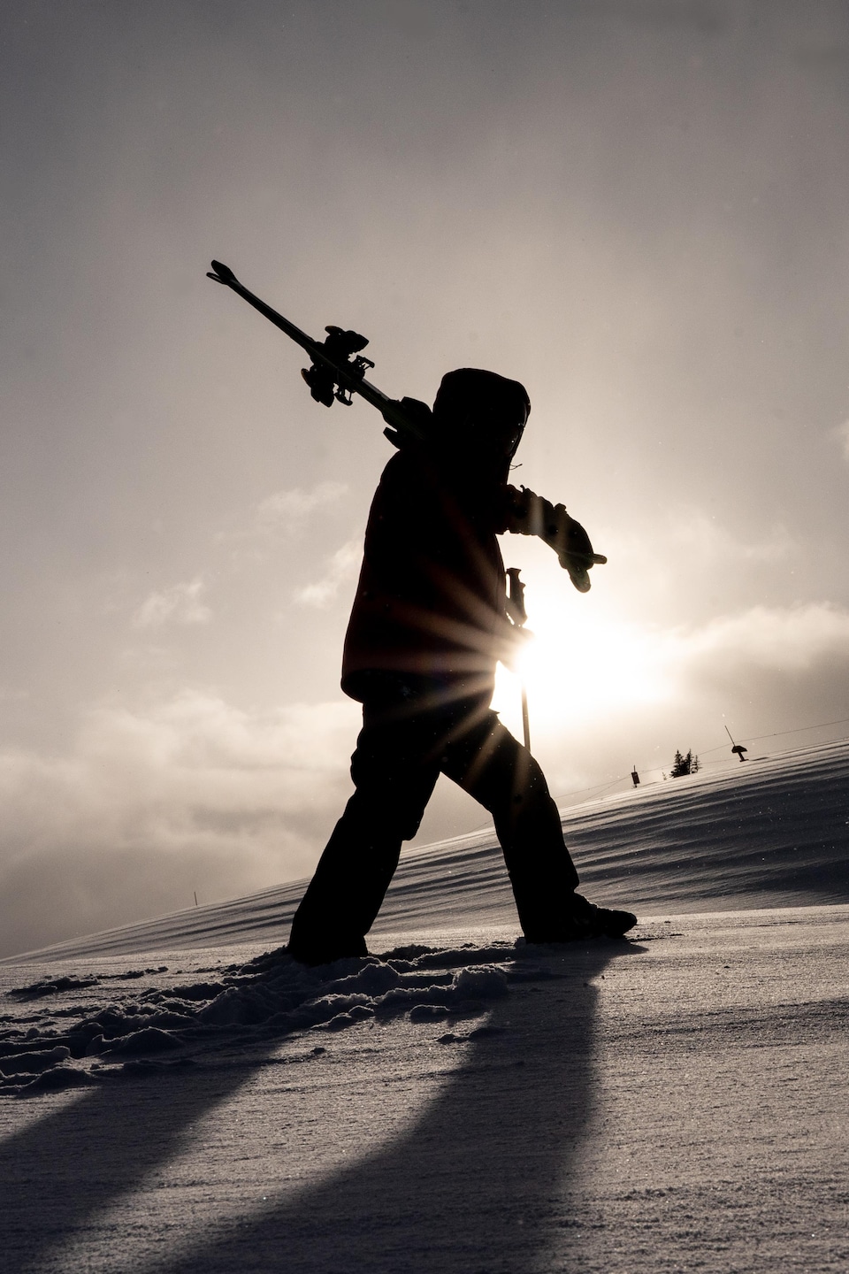 Un homme marche de profil dans la neige devant le soleil avec ses skis dans ses bras.
