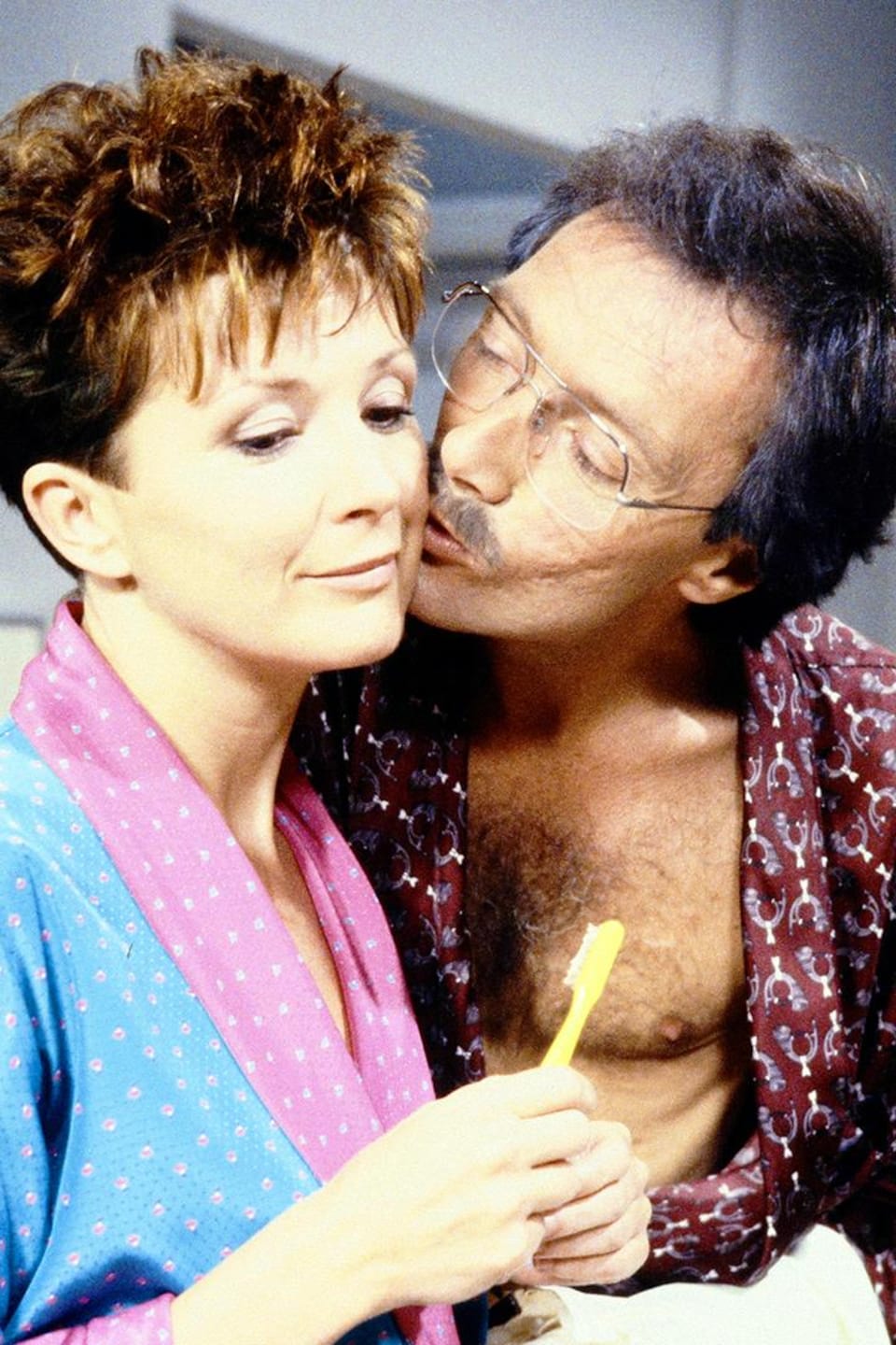 Jean-Paul Belleau (Gilbert Sicotte) embrasse Julie Bastien (Dorothée Berryman) sur la joue. Les deux portent des robes de chambre.