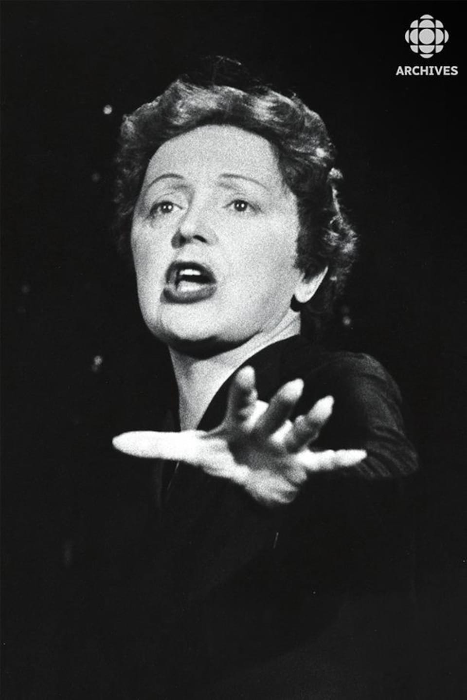 Édith Piaf chante avec une main devant elle. 