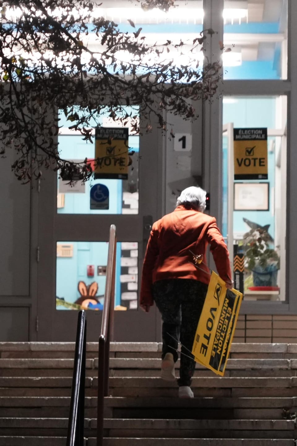 Une dame de dos monte les escaliers en tenant une affiche pour le vote des élections municipales