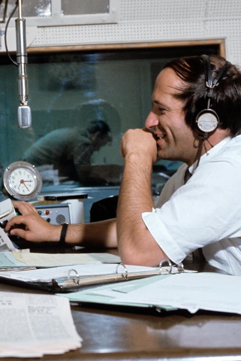 Dans un studio de radio, les animateurs Jean Duceppe et Jacques Houde
discutent, assis de part et d'autre d'un pupitre. Un microphone est suspendu
entre eux. À l'arrière-plan, on voit la régie derrière une vitre.