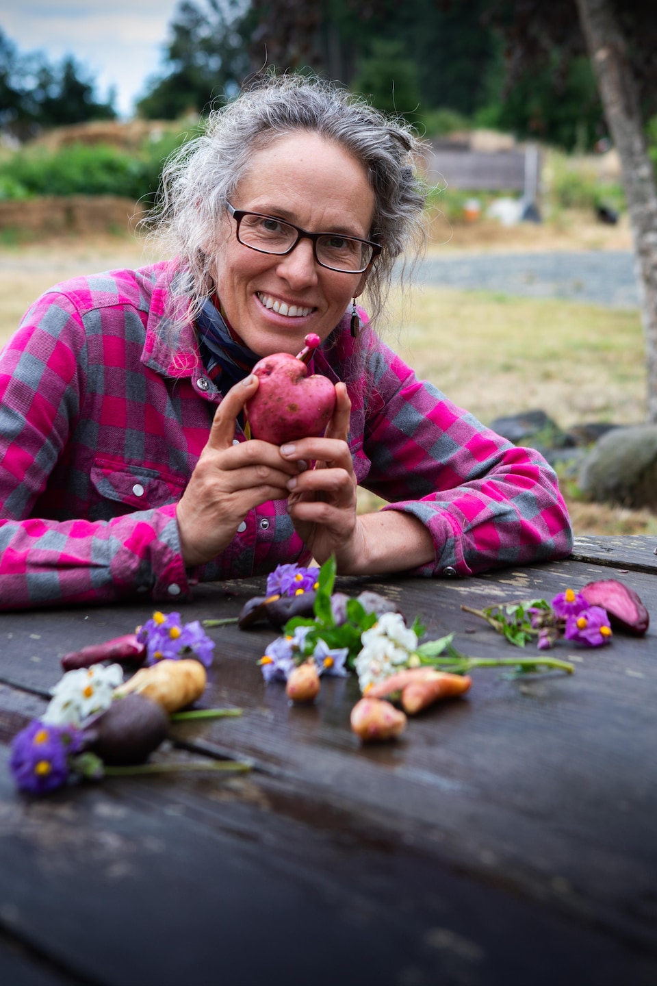 Fiona Hamersley assise à une table en bois avec des fleurs posée dessus, tenant entre ses mains une pomme de terre à la forme de cœur, à la ferme Metchosin, dans le sud de l'île de Vancouver, en août 2024.