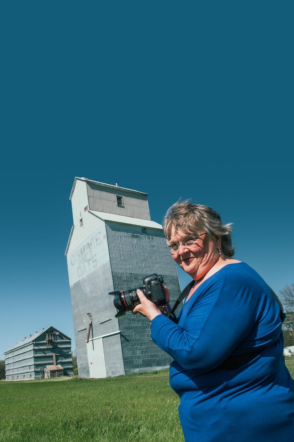 Jean McManus est devant un ancien silo à grain, dans les prairies, elle s'apprête à le prendre en photo avec son appareil photo dans les mains à Snowflake au Manitoba, début mai 2022. 
