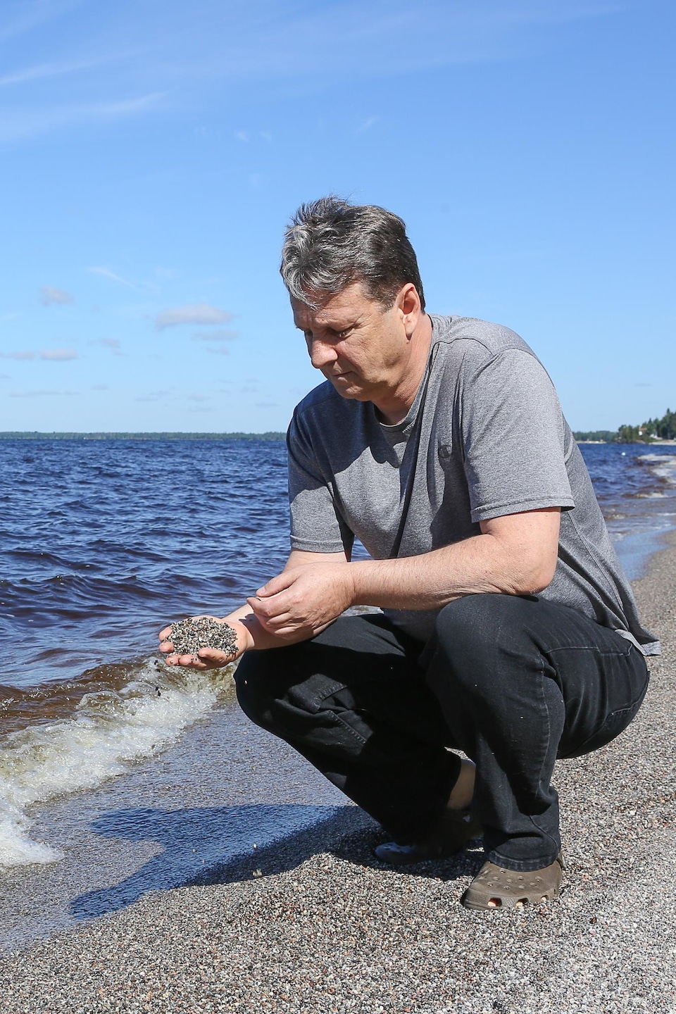 Un homme agenouillé sur la rive d'un lac fait couler du gravillon de sa main.