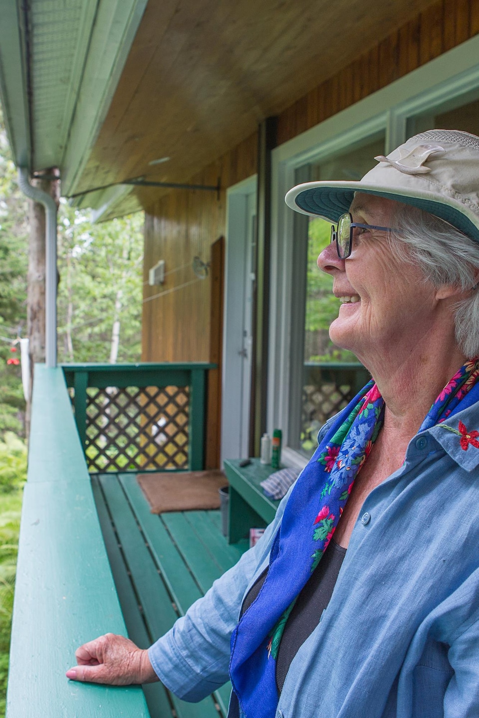 Une femme âgée debout sur la galerie de son chalet regarde vers la forêt.