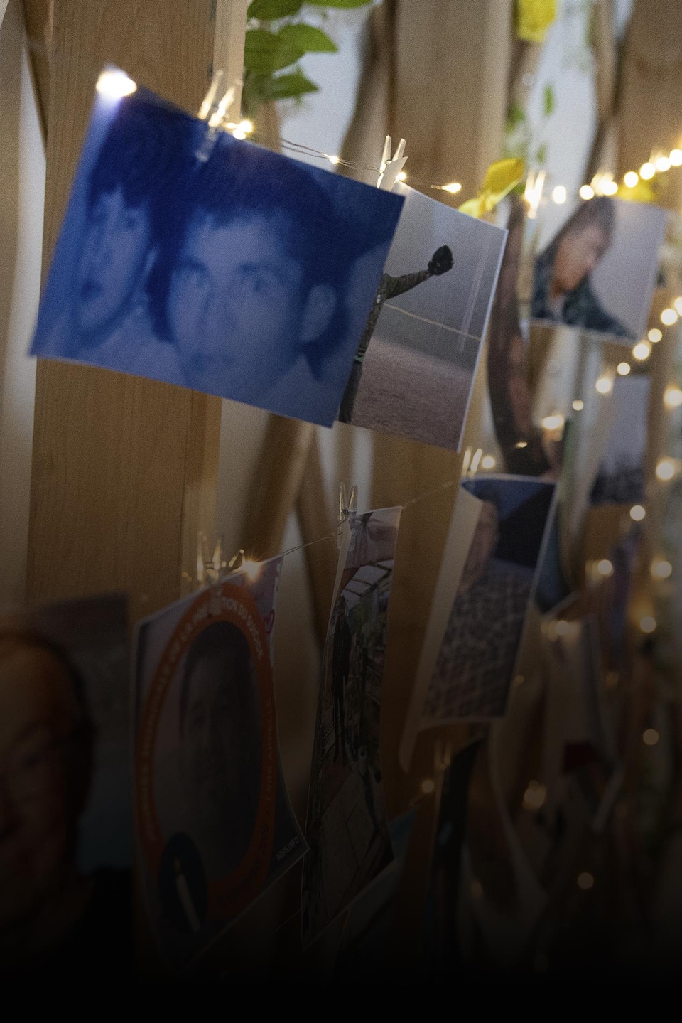 Des photos et portraits de diverses personnes sont accrochés à une structure de bois décorée de petites lumières suspendues.