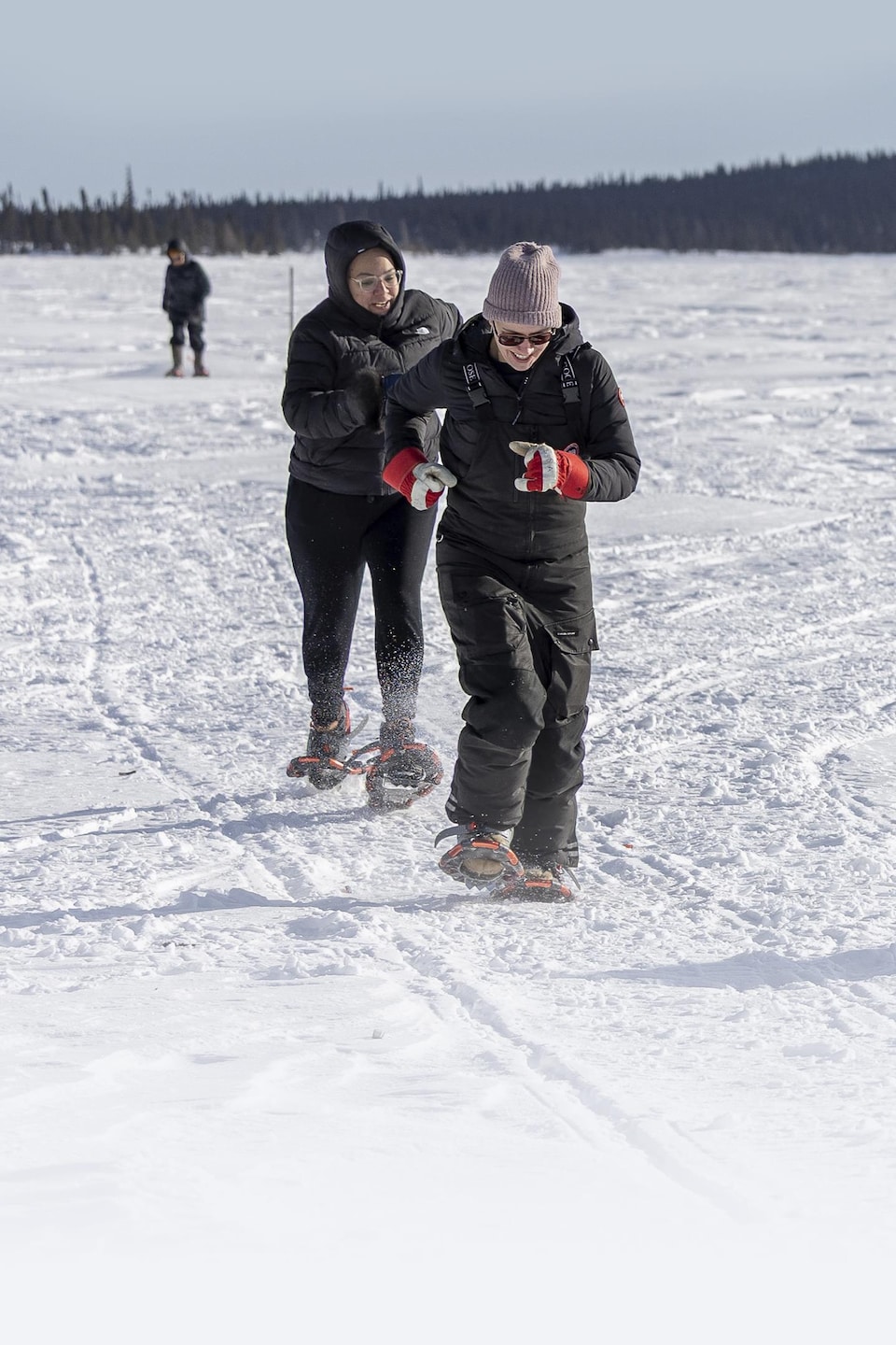 Des personnes participent à une course de raquettes sur une vaste étendue de neige, devant un paysage de collines boisées.