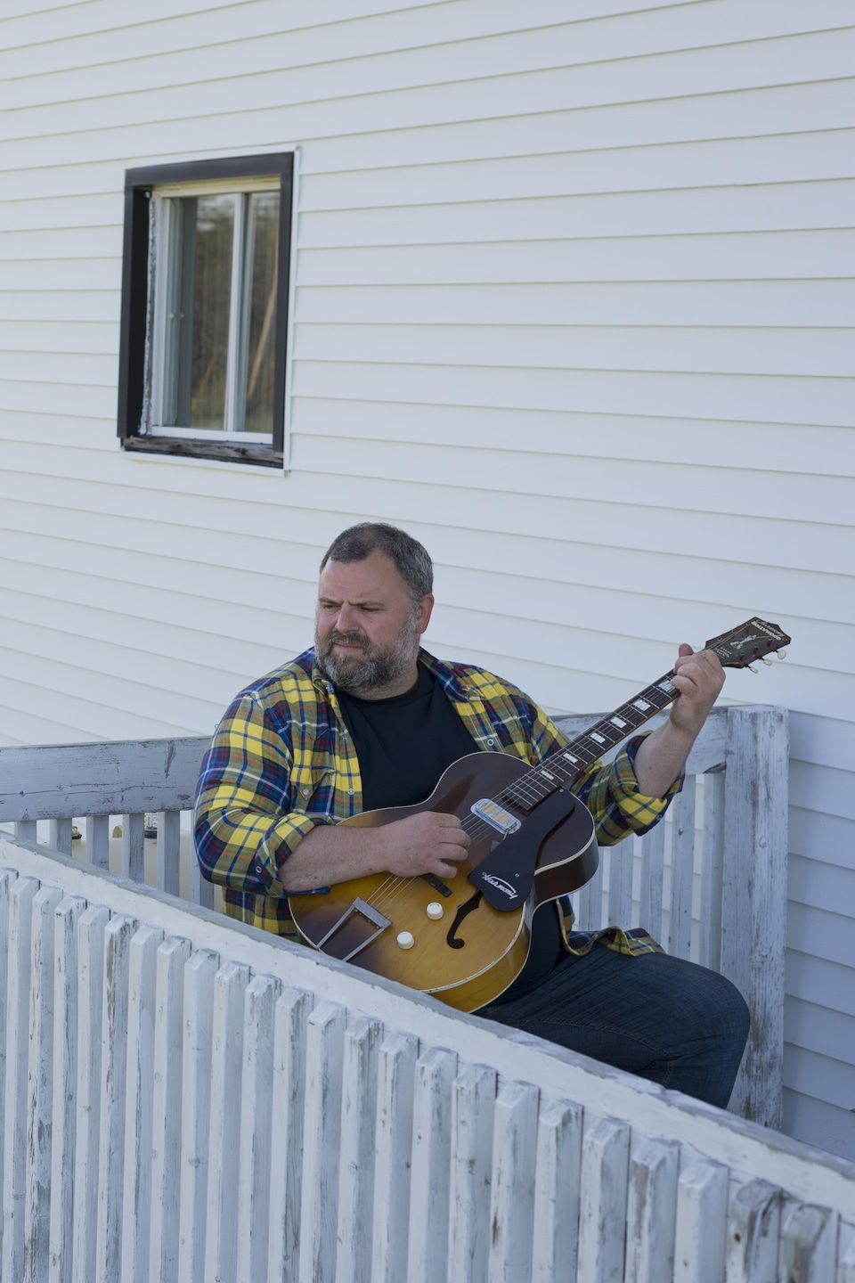 Un homme joue de la guitare assis sur le perron de sa maison blanche.