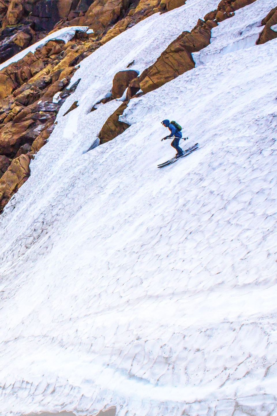Un skieur alpin descend une piste avec peu de neige, entourée de rochers. 