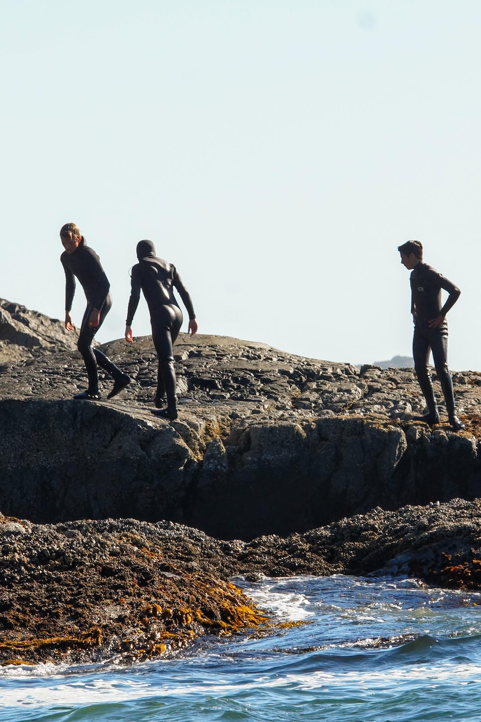 Trois surfeurs en combinaisons hydrofuges sur un rocher près de l'eau.