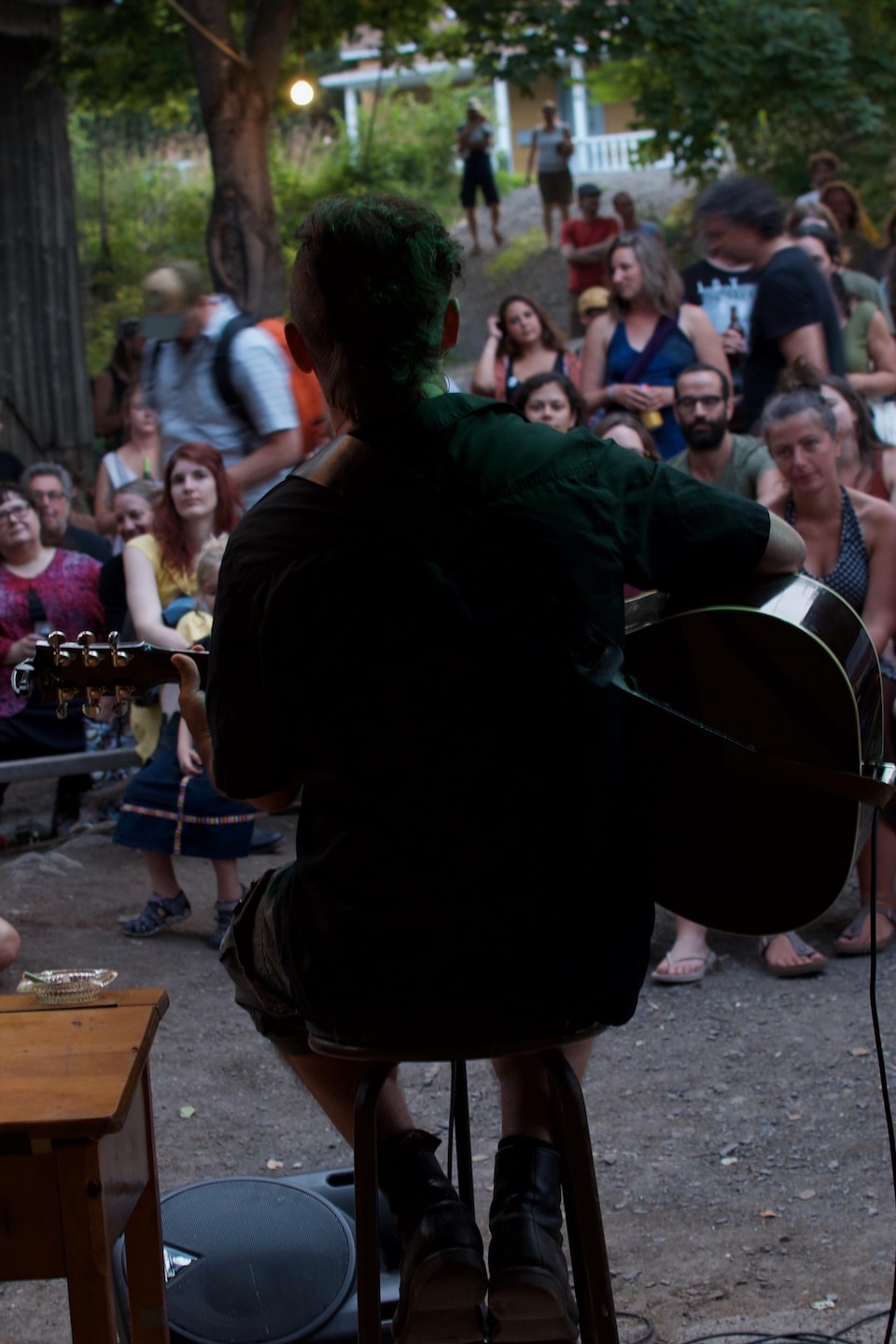 Keith Kouna de dos assis une guitare à la main devant des spectateurs en plein air. 