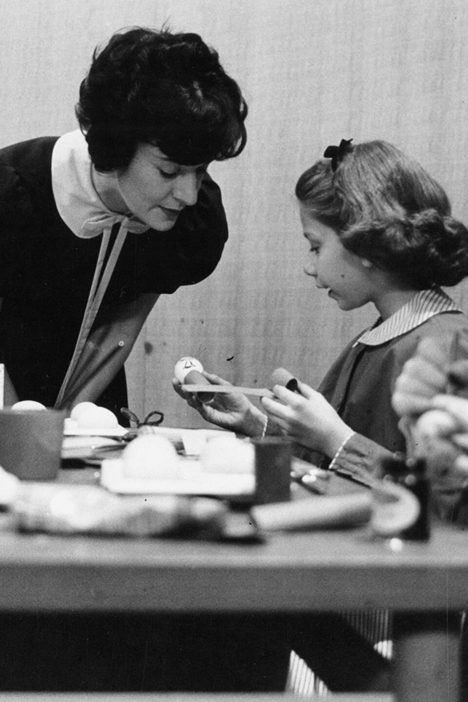 Madeleine Arbour se penche sur le travail d’une jeune fille, attablée devant un bricolage avec un garçon.