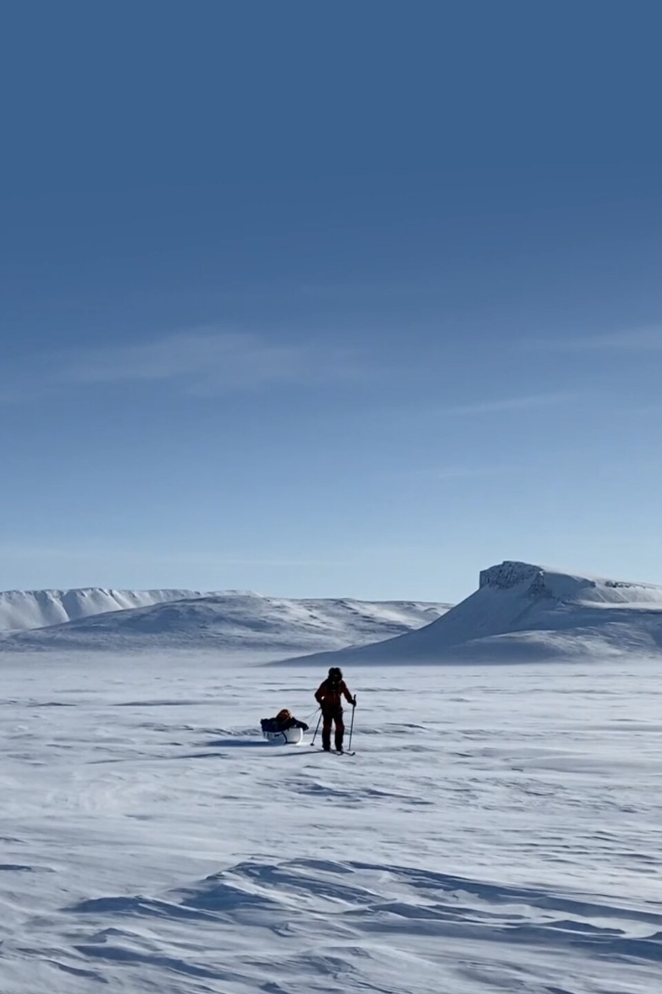 Un aventurier s'avance dans une vaste étendue de neige, skis aux pieds, en traînant derrière lui un traîneau plein de matériel