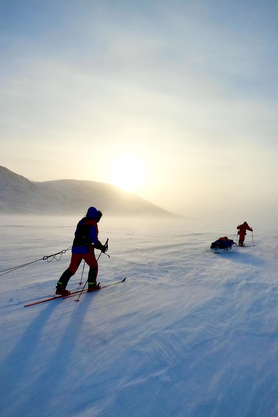 Deux hommes, habillés en épais vêtements d'hiver, skis en traînant un traîneau derrière eux.  Le climat semble froid et aride, alors que la poudrerie souffle sur la neige.