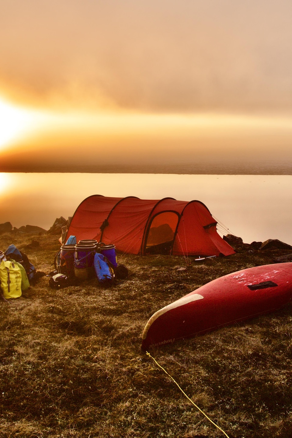 Des sacs à dos, des tonneaux à nourriture, un canot et une grande tente sont installés sur le bord d'un lac, à la levée du jour.
