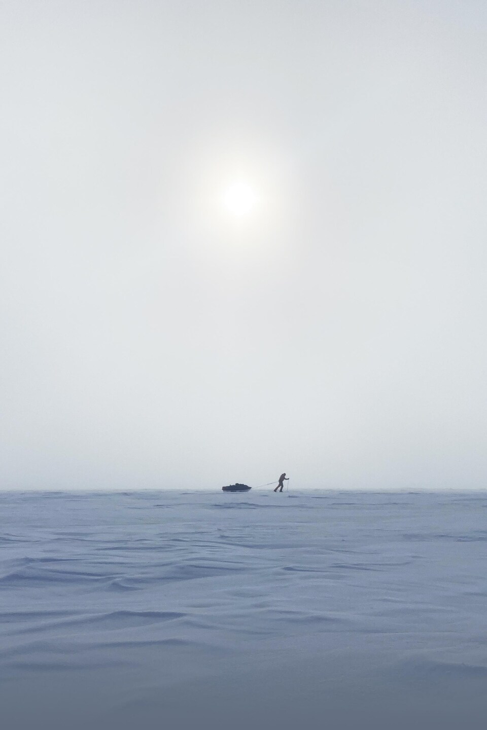 Un homme tirant son traîneau, en ski de fond, avance dans la neige et la poudrerie. Le soleil est caché derrière des nuages.
