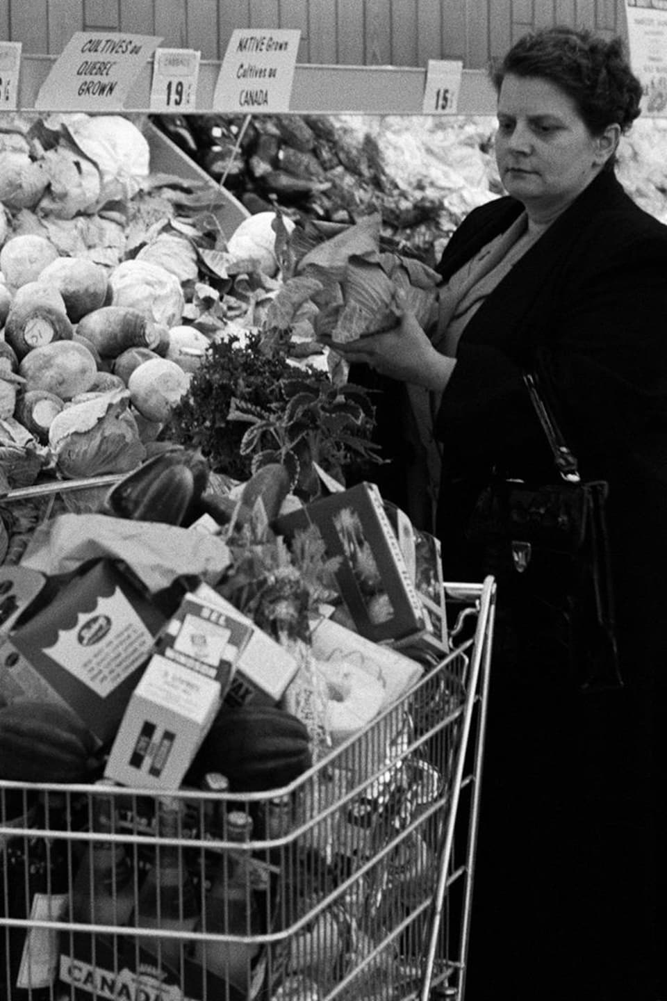 Dans une épicerie, devant le rayon des légumes, une femme est derrière deux paniers remplis d'aliments.