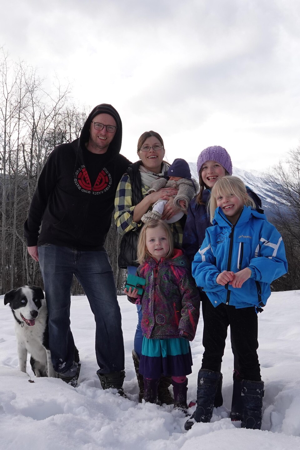 Un père et une mère pose avec leurs trois jeunes filles et leur bébé garçon ainsi que leur chien dans leur cour extérieur enneigée. 