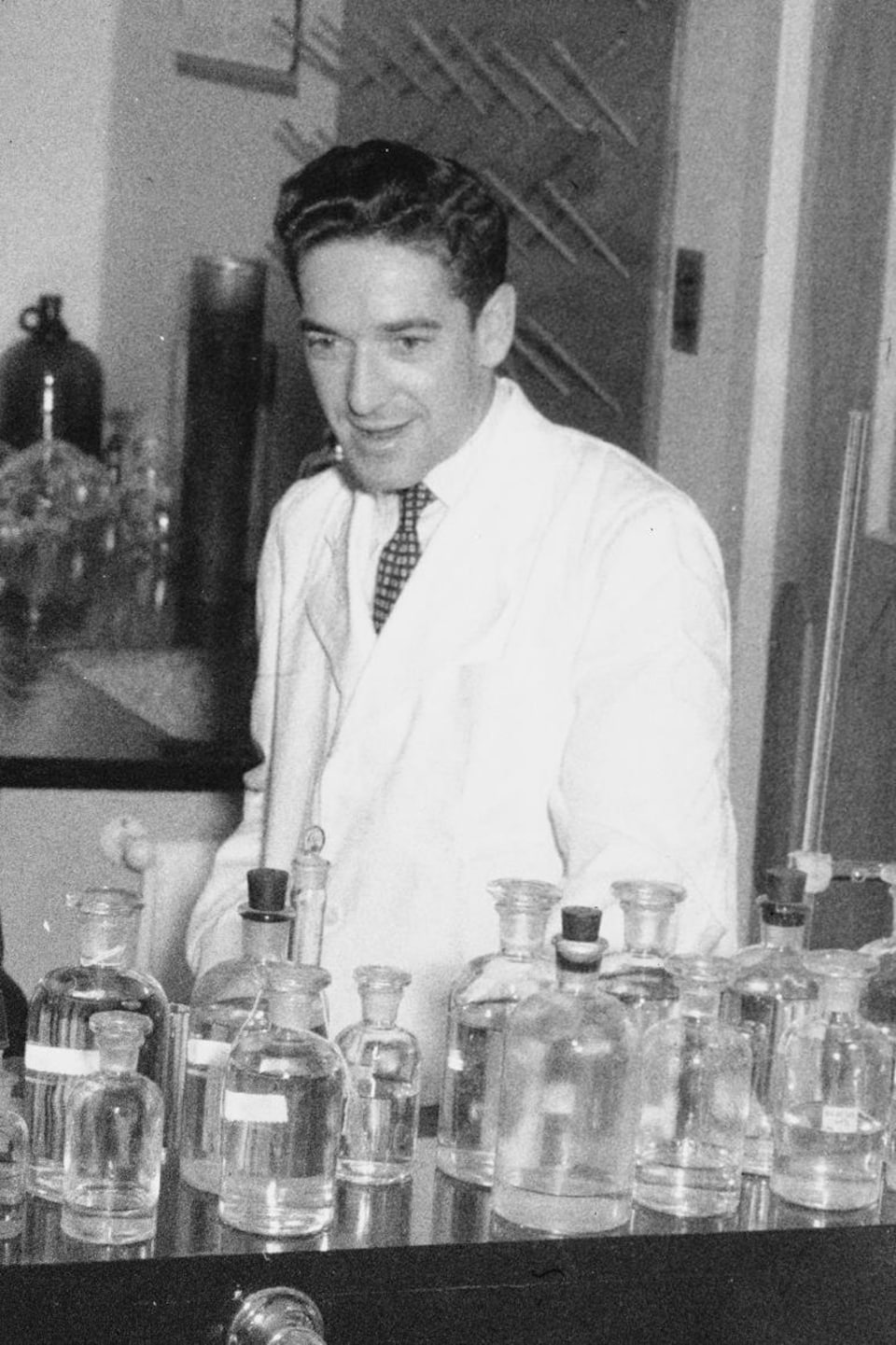 Dans un laboratoire, l'animateur Fernand Seguin derrière une table remplie de fioles. À ses côtés, le Dr Paul Dumas.