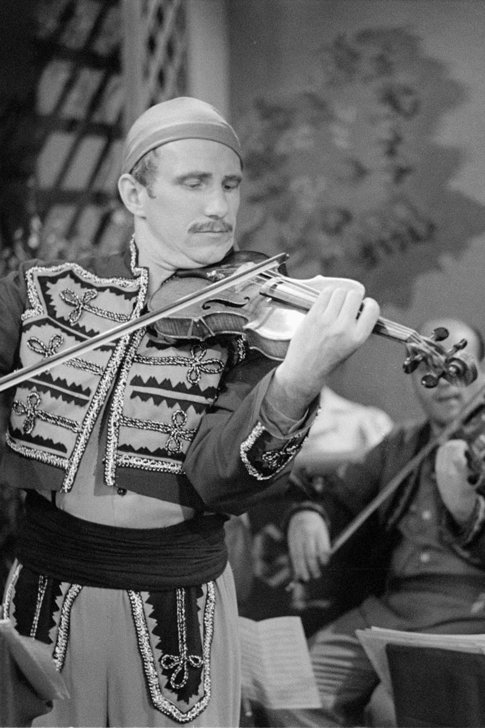 Georges Lapenson joue du violon devant un orchestre en costume folklorique.