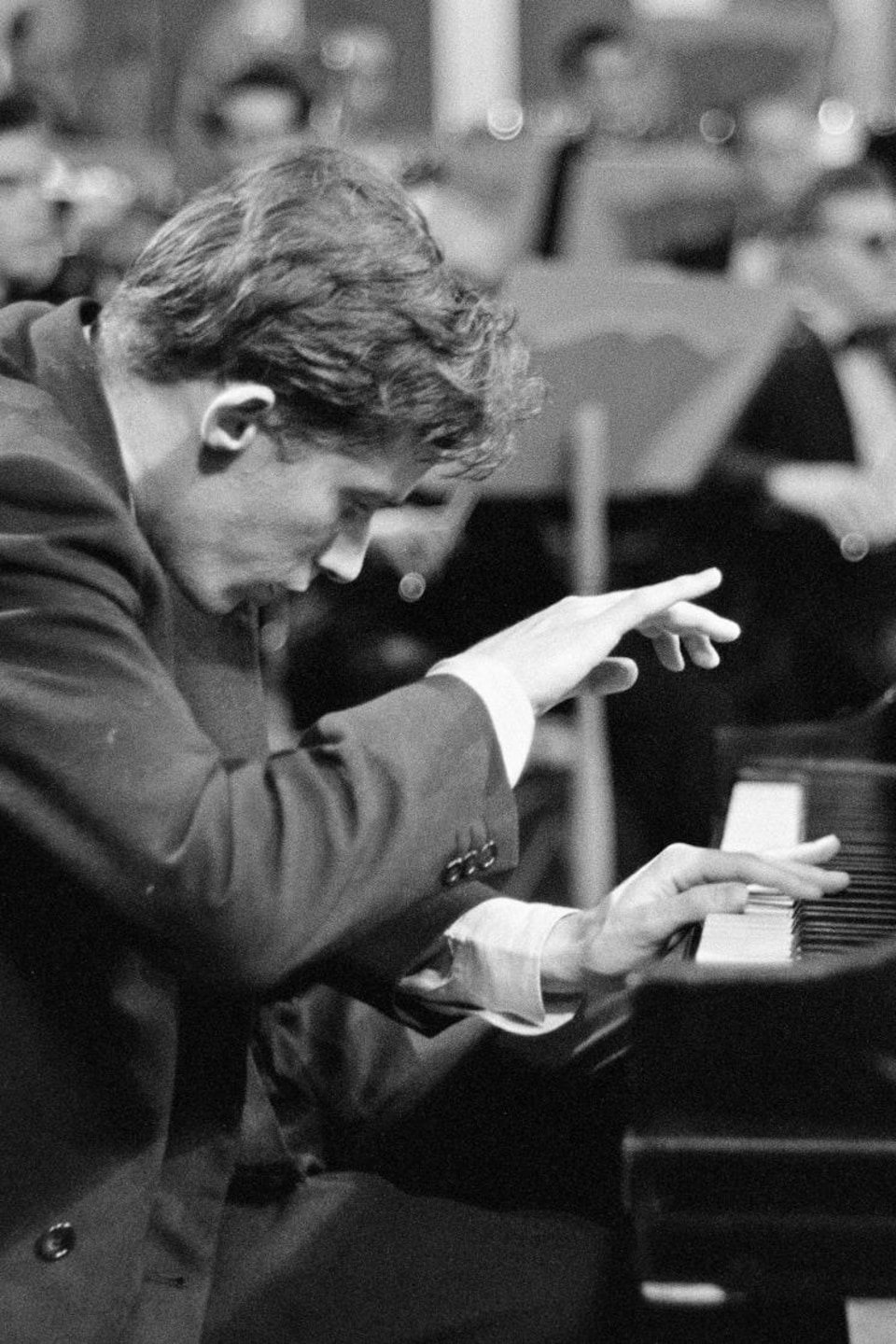 Glenn Gould en performance au piano, les mains en mouvement au-dessus de son instrument avec un orchestre en arrière-plan.
